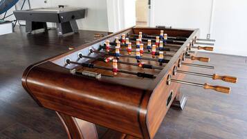 Une table de baby-foot dans une pièce au plancher en bois, avec une table de billard en arrière-plan.