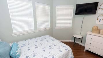 Un dormitorio con cama, dos ventanas con persianas, un televisor en la pared, un tocador pequeño y blanco, y una mesa redonda.