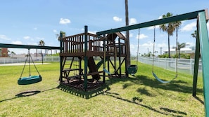 A playground with swings and a wooden climbing structure. - Family Condo - Pool, Jacuzzi & BBQ - Walk to Beach (South Padre Island)