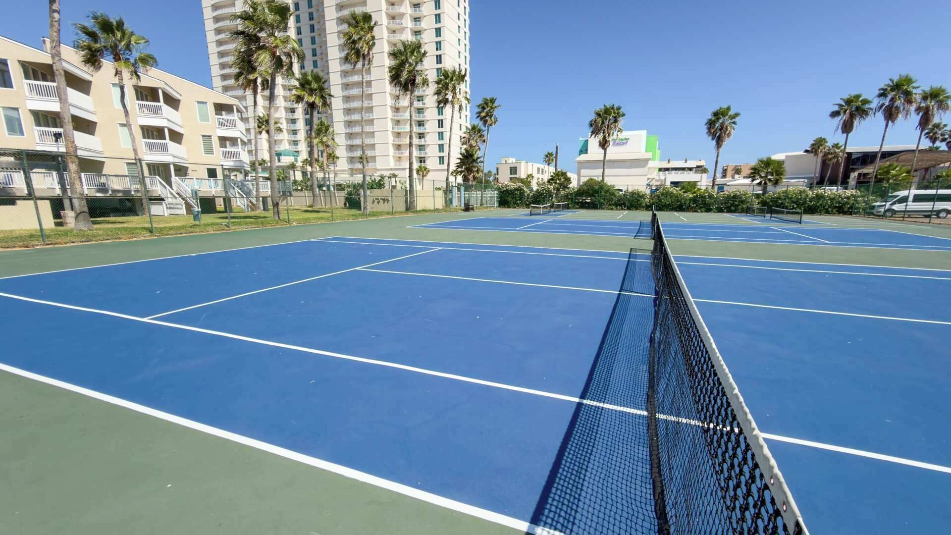 A tennis court with a net in the foreground, surrounded by palm trees and buildings.