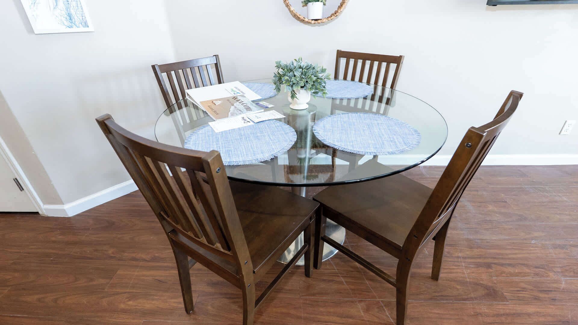 Una mesa de vidrio redonda con cuatro sillas de madera, una pequeña planta en maceta y manteles individuales.