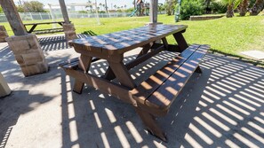 Una mesa de picnic con sombra, rodeada por una superficie de hormigón y un área de césped.