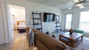 A modern living room with a sofa, a coffee table, a TV mounted on the wall, and a balcony visible through the window. - Beachfront 2br, 5th Floor Views, Walkable SPI (South Padre Island)