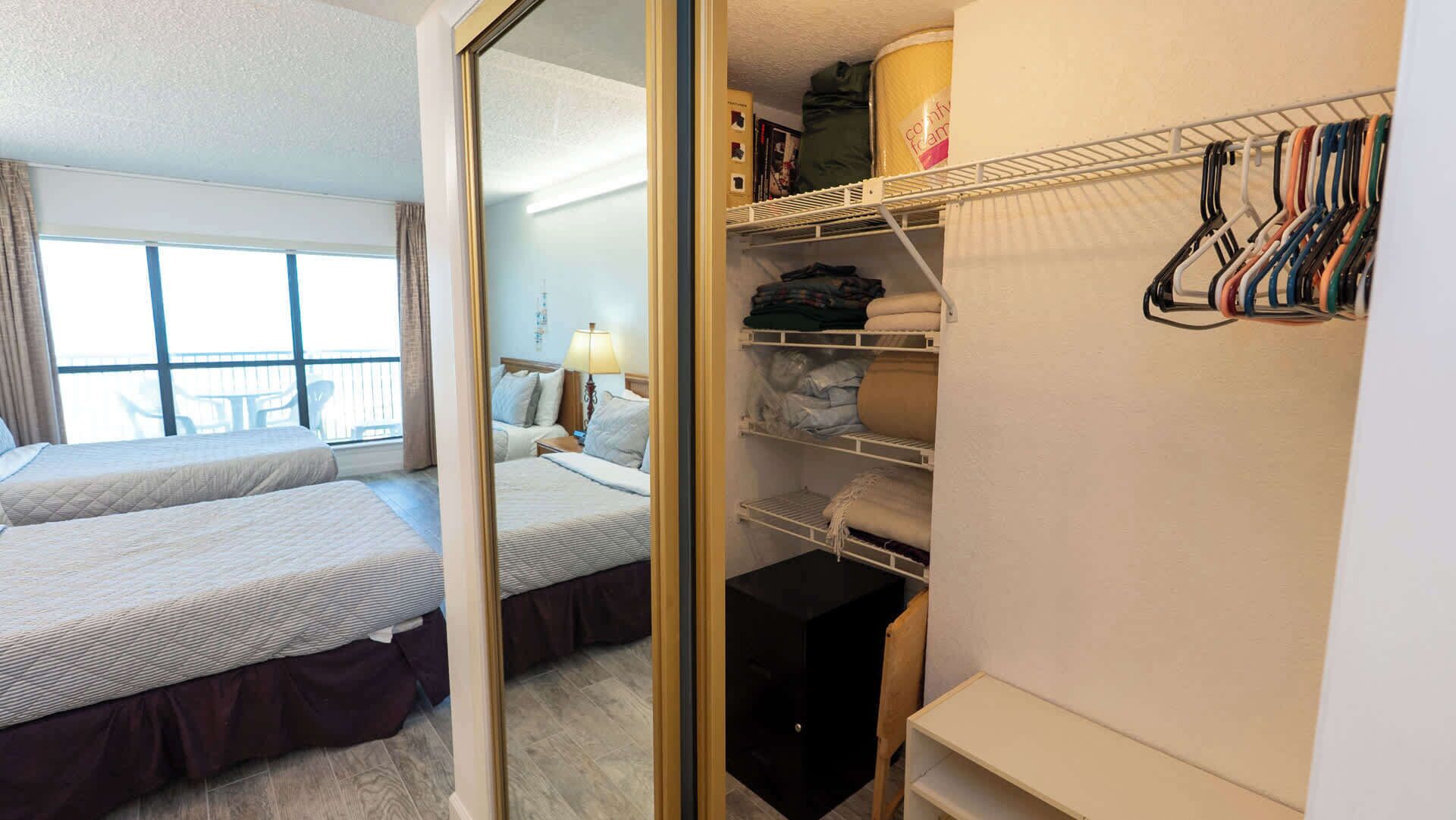 Quarto de hotel com duas camas, um guarda-roupa com roupas penduradas e vista para o exterior.