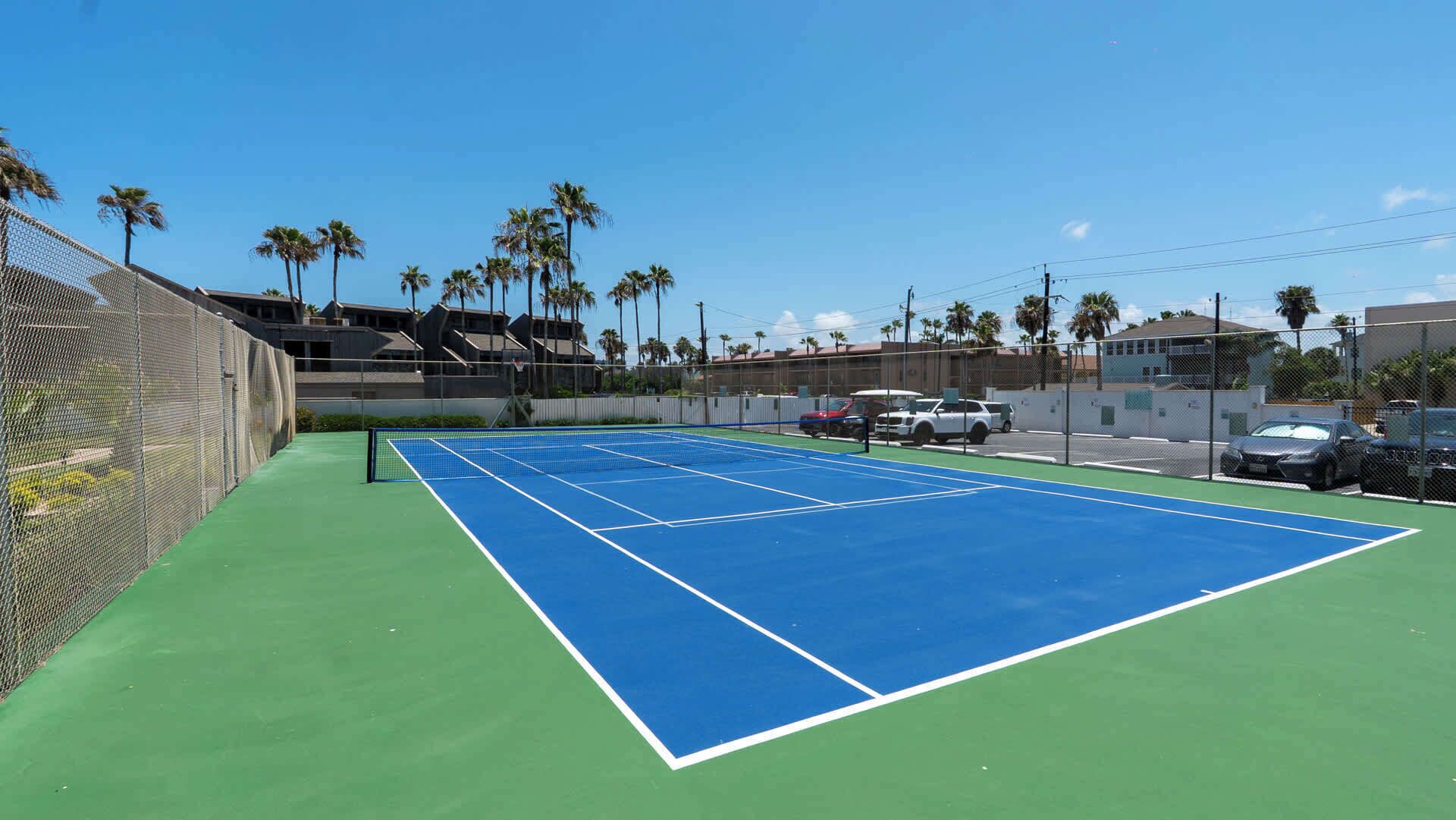 An outdoor tennis court with a green and blue surface, surrounded by a fence and palm trees.