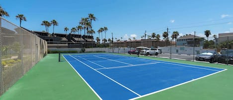 An outdoor tennis court with a green and blue surface, surrounded by a fence and palm trees.