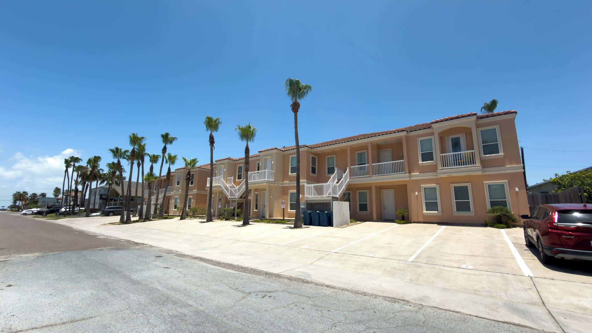 A multi-story residential building with balconies, a parking lot, and palm trees.