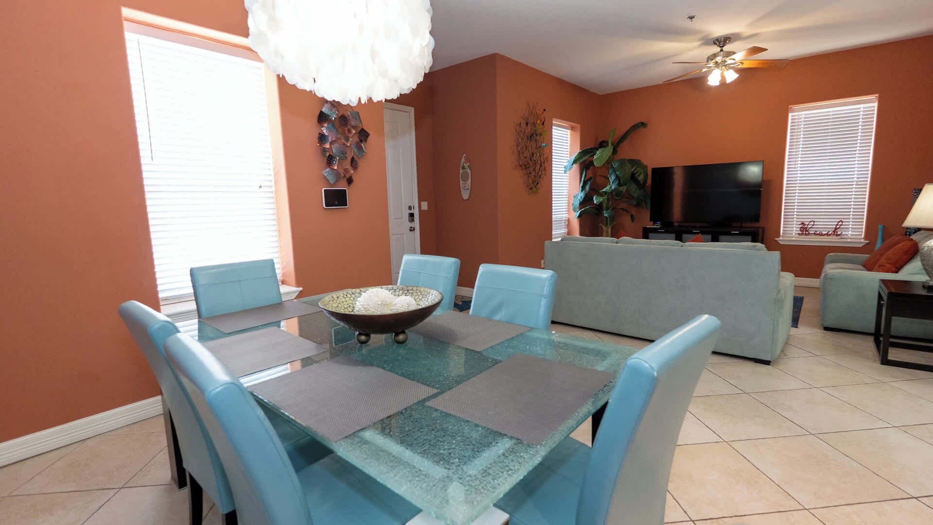 A living room with a glass-top dining table, blue chairs, a sofa, a television, and a ceiling fan.