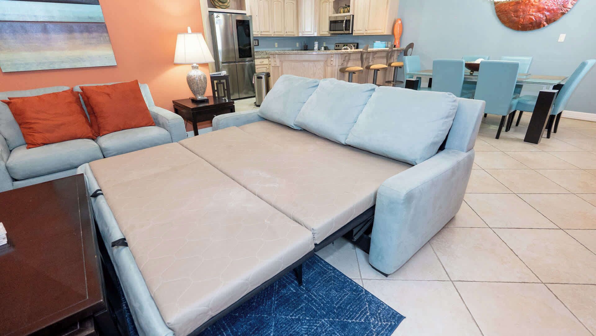 A convertible sofa bed in a living room with a kitchen in the background.