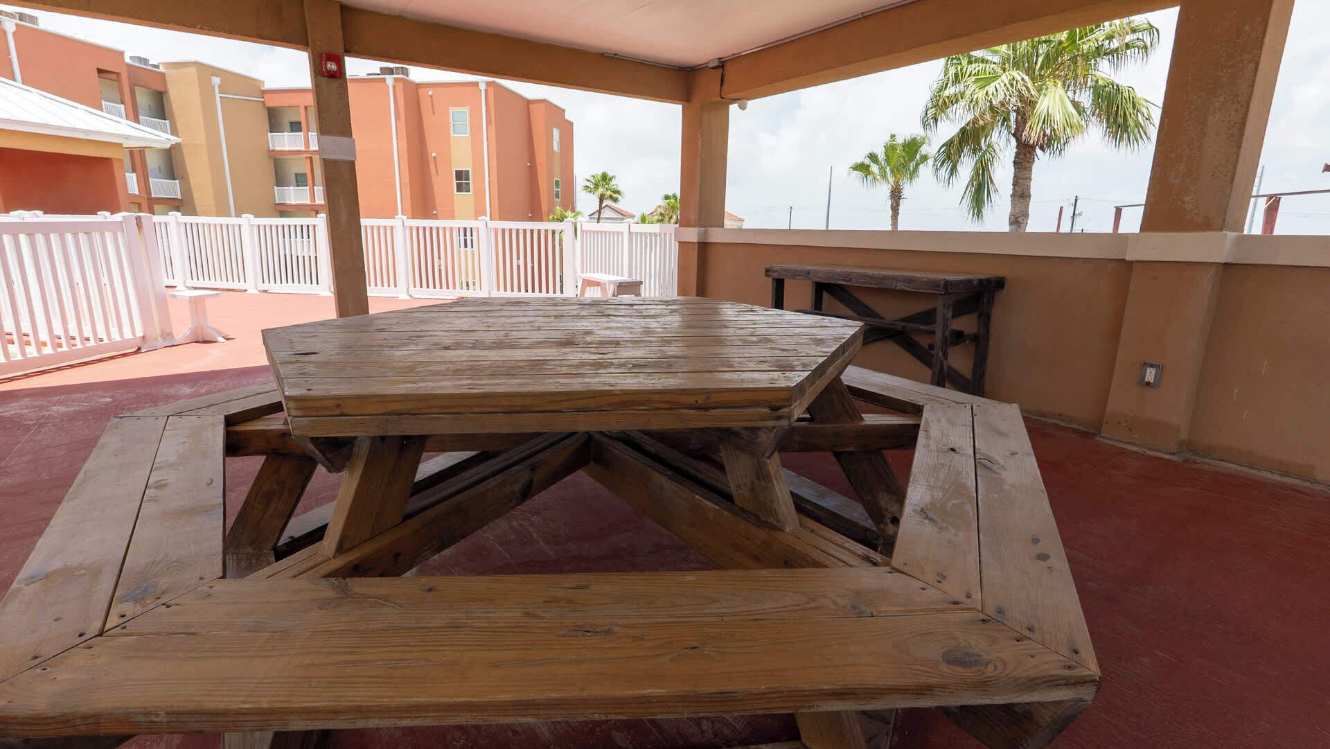 Taula de picnic de fusta en una zona exterior coberta amb vista a un edifici i palmeres.