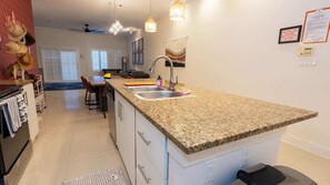 A modern kitchen with granite countertops, a built-in dishwasher, and a large island with a sink. There is a dining area with a table and chairs, and a ceiling fan. - 3BR Condo - 9 Beds - Pool - Walk to Beach - SPI (South Padre Island)