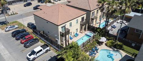 A two-story building with a swimming pool, a parking lot with several cars, and a small garden area with palm trees.