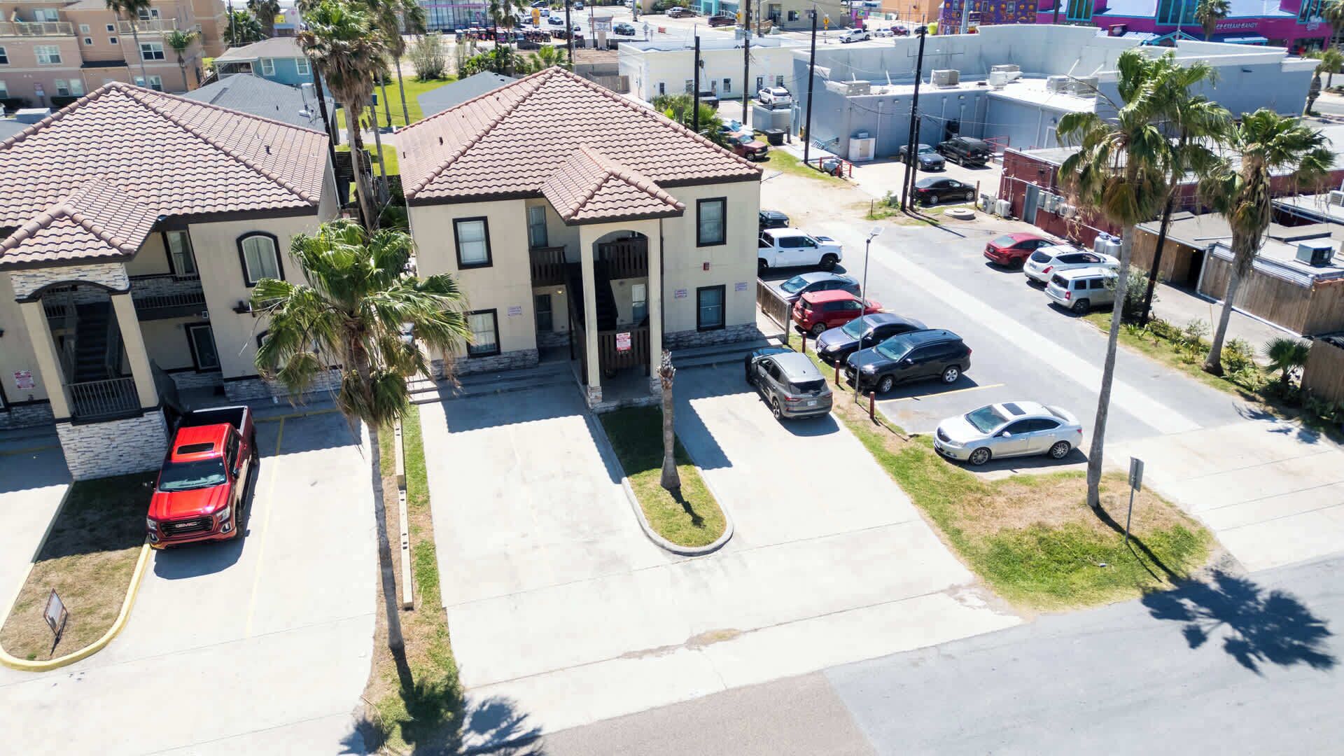 A multi-story building with a parking lot, palm trees, and a red vehicle parked in front.