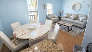 A dining area with a round table and chairs, a living room with a sofa and coffee table, and a wall with decorative items. - Near Beach Retreat - Ground Floor - Pool - Walk to All (South Padre Island)