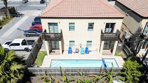 Ein zweistöckiges Gebäude mit Swimmingpool, einem Balkon mit blauen Stühlen und Blick auf eine Straße mit parkenden Autos.
