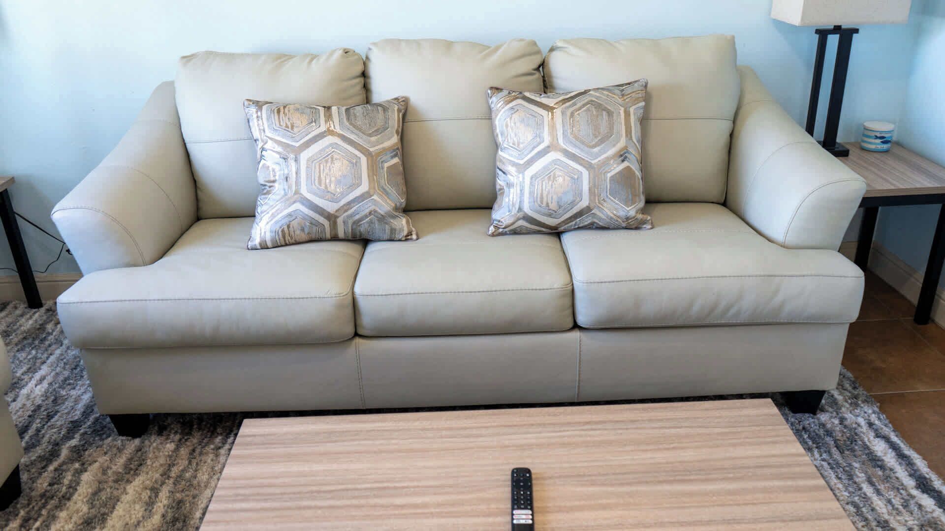 Ein beiges Sofa mit gemusterten Kissen, eine Fernbedienung auf einem Holztisch und eine Lampe auf einem Beistelltisch.