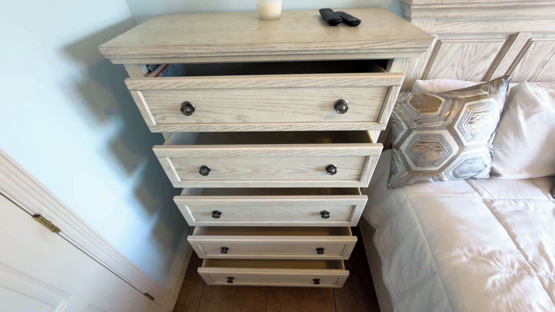 A wooden dresser with multiple drawers and knobs, a remote control on top, and a patterned pillow on the bed.