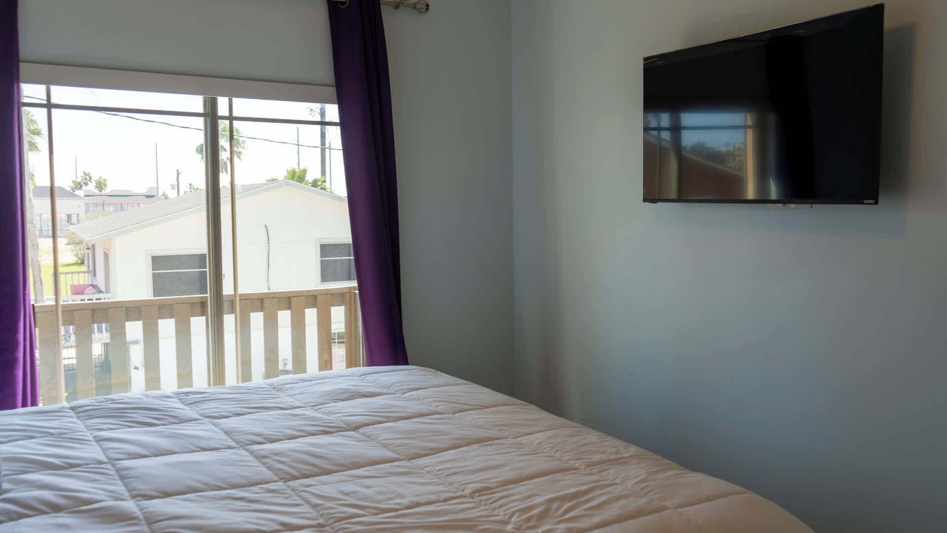 Une chambre à coucher avec un lit, un téléviseur fixé au mur et une fenêtre avec des rideaux.