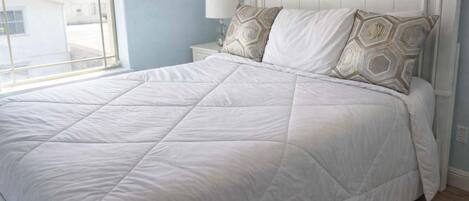 A bed with a white quilt, two patterned pillows, a nightstand with a lamp, and a window with a view of buildings.