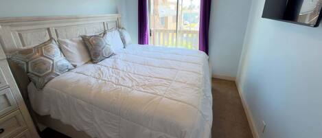 A neatly made bed with a headboard, a window with purple curtains, and a wall-mounted TV.