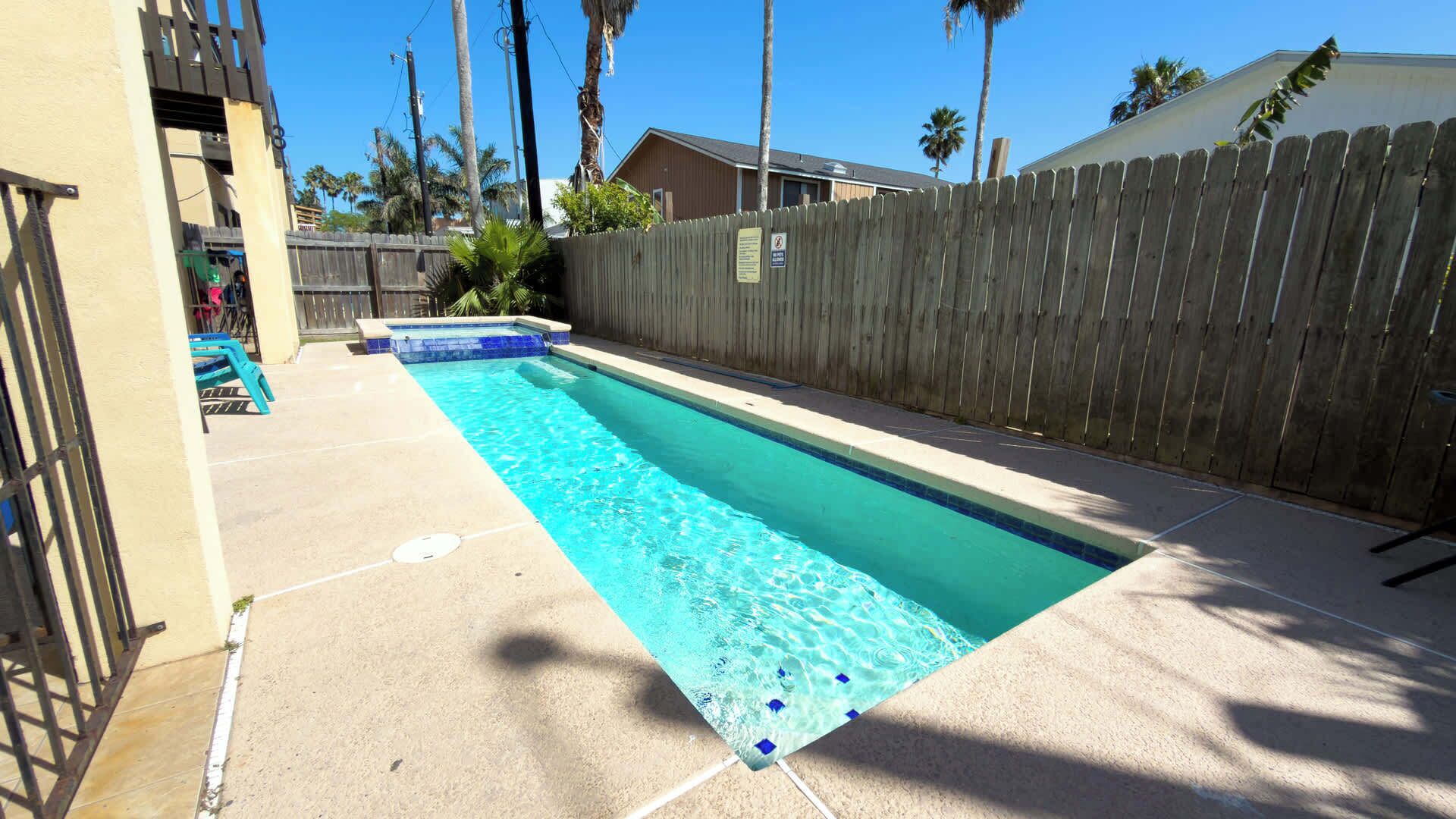 Uma piscina retangular com água azul cristalina, cercada por uma vedação de madeira e um deck de concreto.