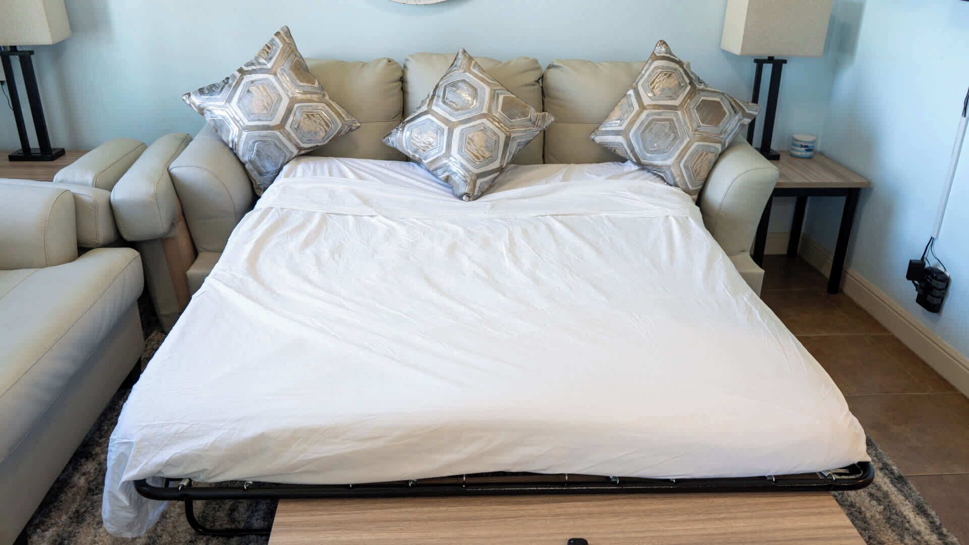 Una cama con colchón blanco y cuatro almohadas estampadas, un sofá color beige, dos mesitas de noche con lámparas y suelo de madera.