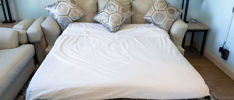 A bed with a white mattress and four patterned pillows, a beige sofa, two nightstands with lamps, and a wooden floor.
