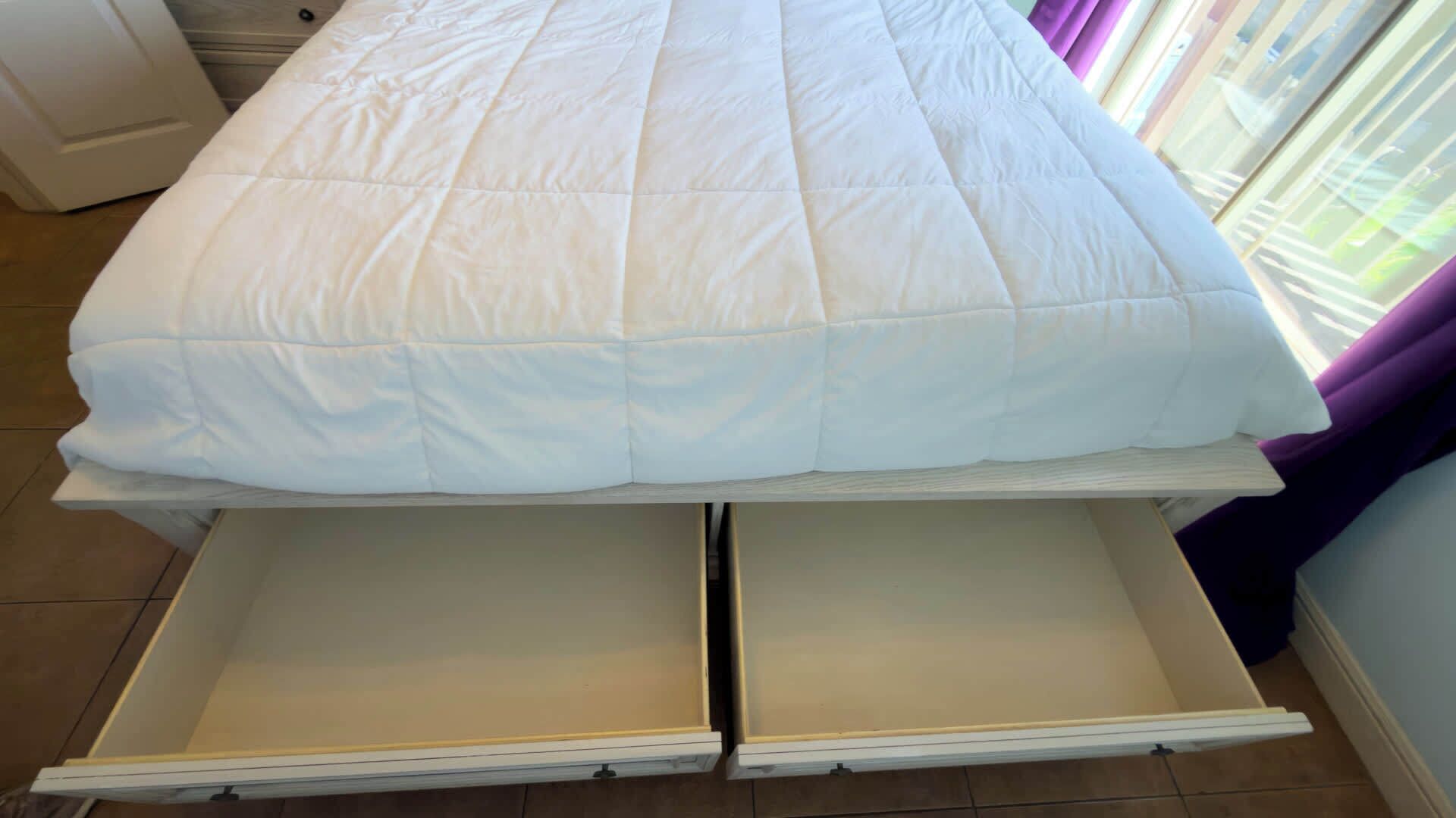 A bed with a white duvet, a drawer below it, and a window with purple curtains.