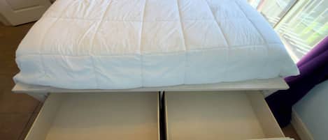 A bed with a white duvet, a drawer below it, and a window with purple curtains.
