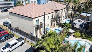 A multi-story building with a swimming pool, a fenced parking area, and a view of the surrounding neighborhood. - Near Beach Retreat - Ground Floor - Pool - Walk to All (South Padre Island)