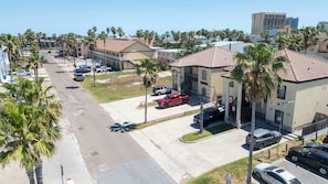 A residential street with parked cars, palm trees, and a building with a tiled roof. - Near Beach Retreat - Ground Floor - Pool - Walk to All (South Padre Island)