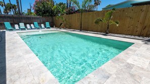 Une piscine rectangulaire à l’eau claire, entourée d’une clôture en bois, et bordée d’une rangée de chaises longues turquoise.