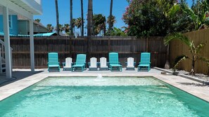 Uma piscina com uma fonte, rodeada por seis cadeiras Adirondack turquesa, um cercado de madeira e palmeiras.