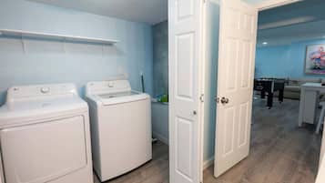 A laundry room with two white washers and dryers, a door leading to a living area with a pool table, and a painting on the wall.
