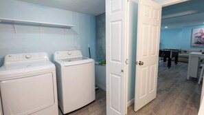 A laundry room with two white washers and dryers, a door leading to a living area with a pool table, and a painting on the wall. - New 5BR Remodel, Heated Pool, Game Room (South Padre Island)