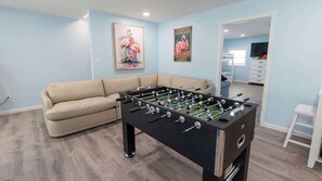 A room with a foosball table, a sofa, and two wall-mounted paintings. - New 5BR Remodel, Heated Pool, Game Room (South Padre Island)