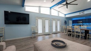 A spacious living room with a flat-screen TV, a dining area with chairs, a kitchen with a microwave, and a ceiling fan.
