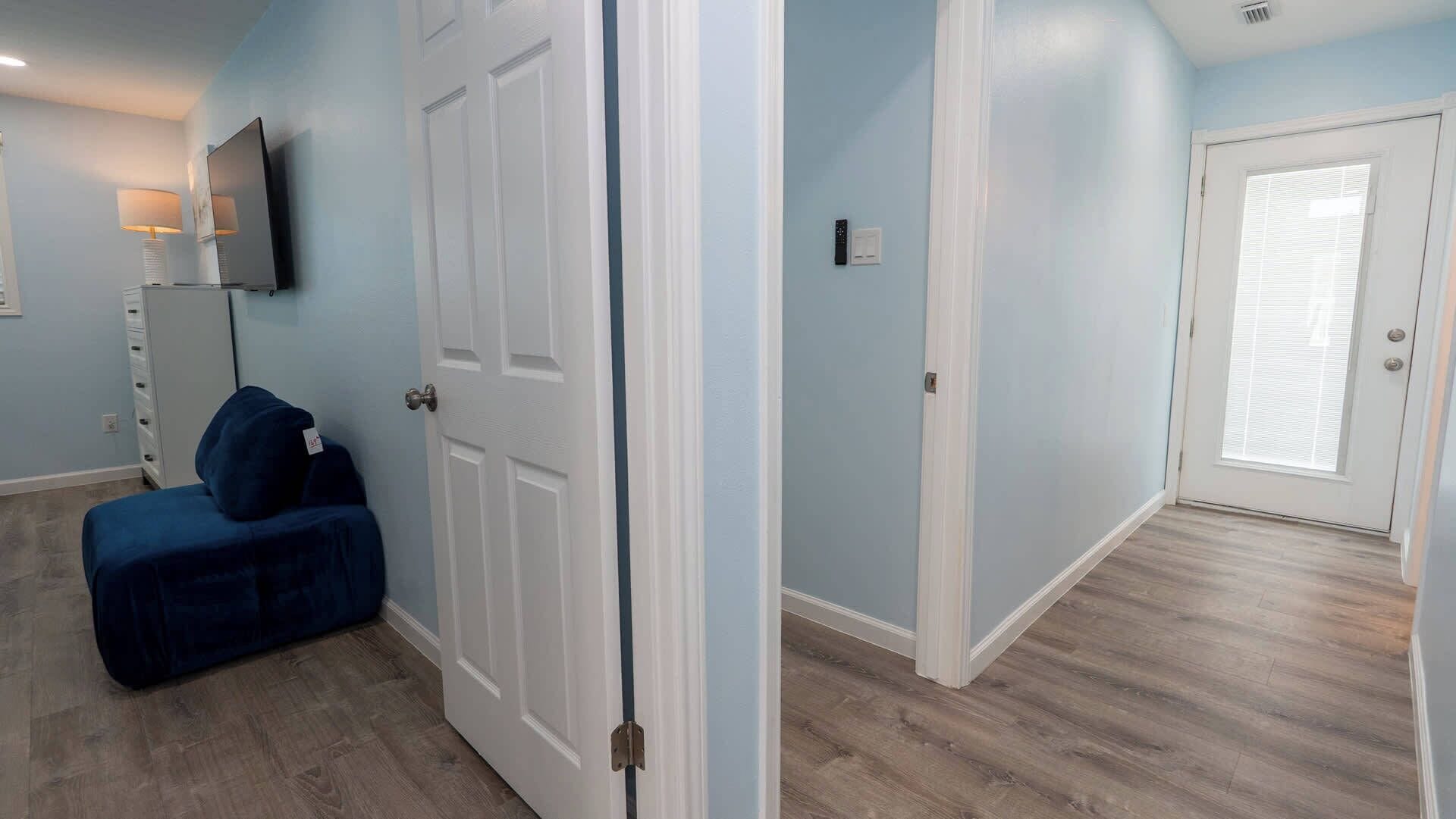 A room with a closed door, a blue ottoman, a TV on the wall, and a window with blinds.