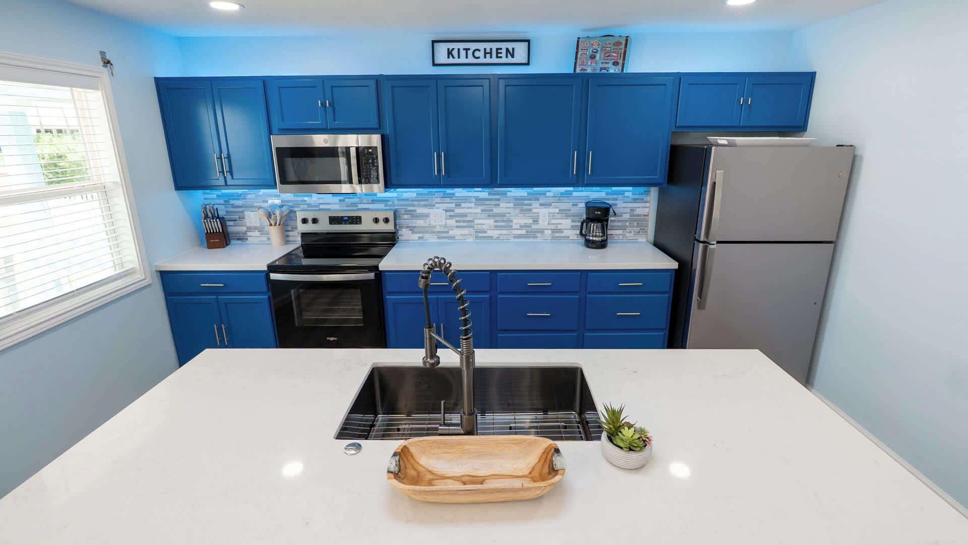 A modern kitchen with blue cabinets, stainless steel appliances, a white countertop, and a wooden serving tray.