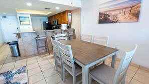 Un comedor con una mesa y sillas de madera, una cocina al fondo y un cuadro de una playa en la pared.