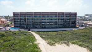 Un hotel de varios pisos con piscina, rodeado de vegetación y una zona de arena.