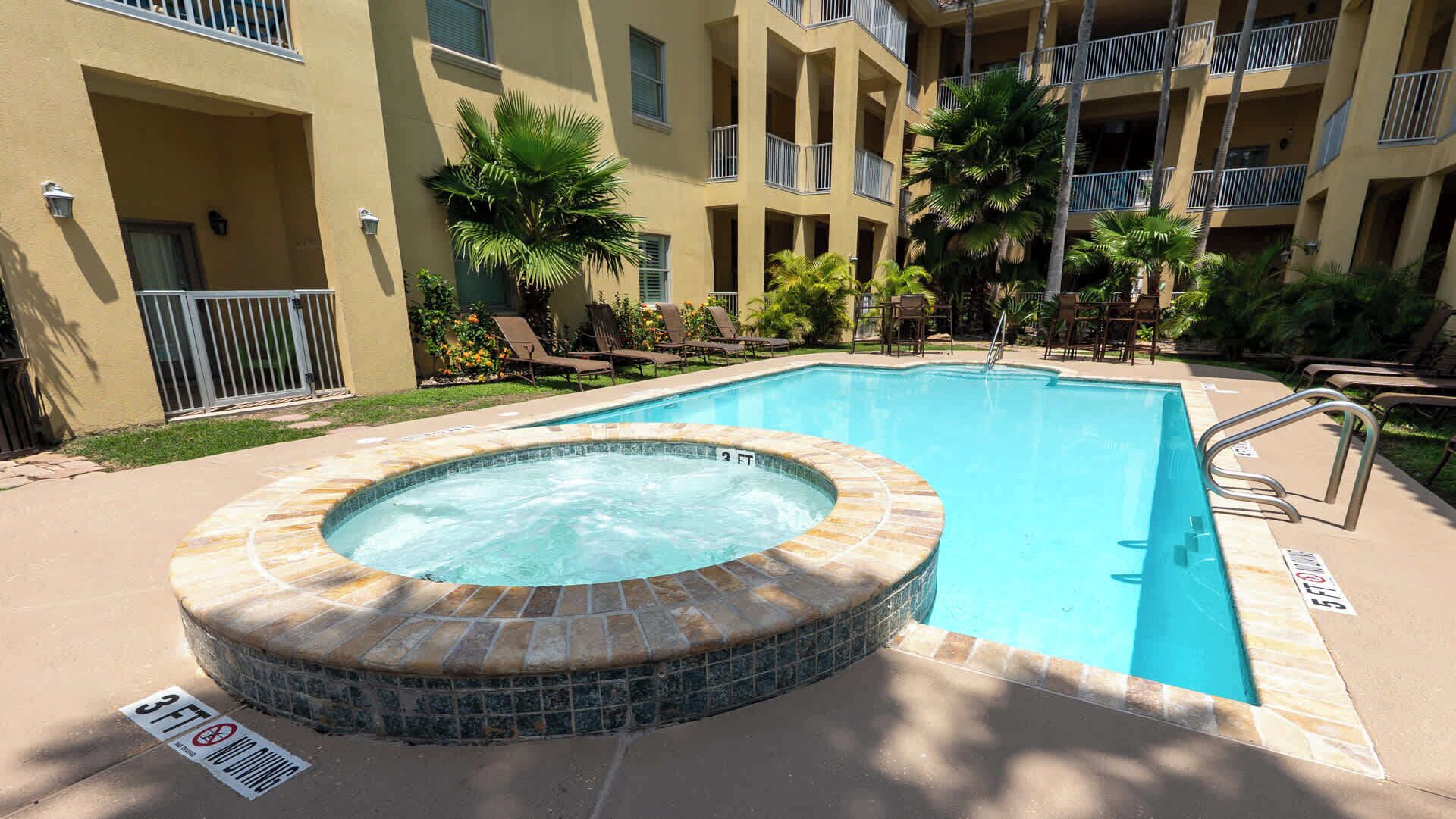 A swimming pool with a hot tub, surrounded by lounge chairs and palm trees.