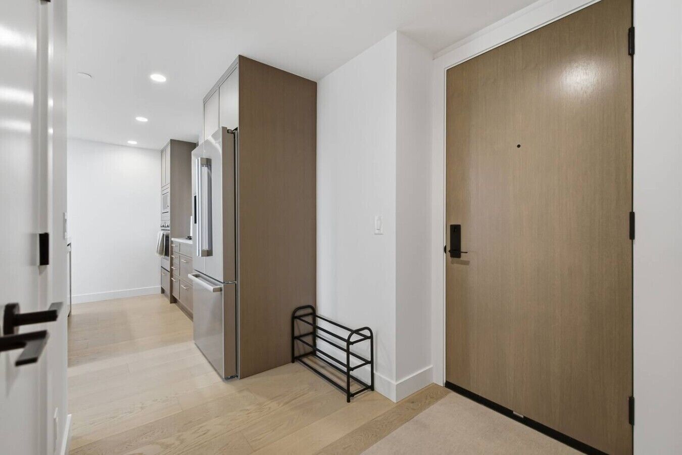 Un couloir avec une porte en bois, un étagère en métal et un coin cuisine équipé de placards et d’appareils électroménagers.