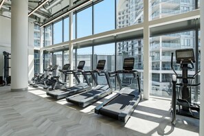 A gym with treadmills and exercise equipment, large windows, and a view of high-rise buildings. - Luxury Beachfront | 2 Bed | Pool & Hot Tub | Gym (Kelowna)