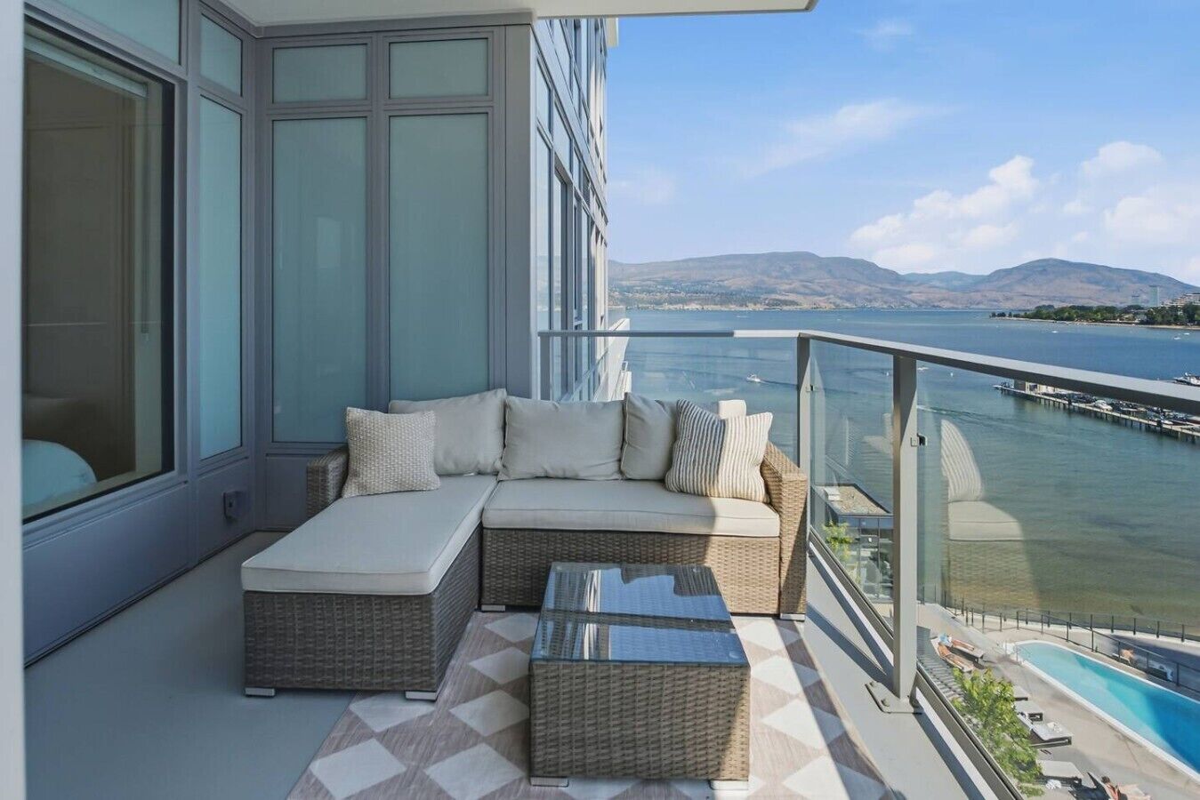 Ein Balkon mit einer Couch, einem Glastisch und Blick auf ein Gewässer und weit entfernte Hügel.
