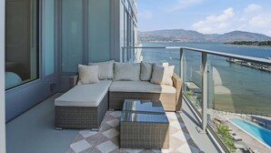 A balcony with a sofa, a glass coffee table, and a view of a body of water and distant hills. - Luxury Beachfront | 2 Bed | Pool & Hot Tub | Gym (Kelowna)
