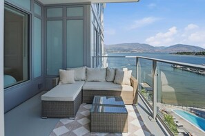 A balcony with a sofa, a glass coffee table, and a view of a body of water and distant hills. - Luxury Beachfront | 2 Bed | Pool & Hot Tub | Gym (Kelowna)