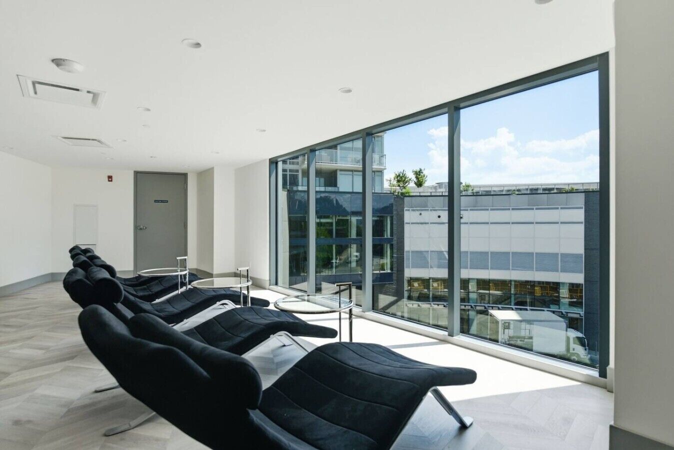 Un espace d’attente moderne avec des chaises noires, un mur de verre et une vue sur un immeuble.