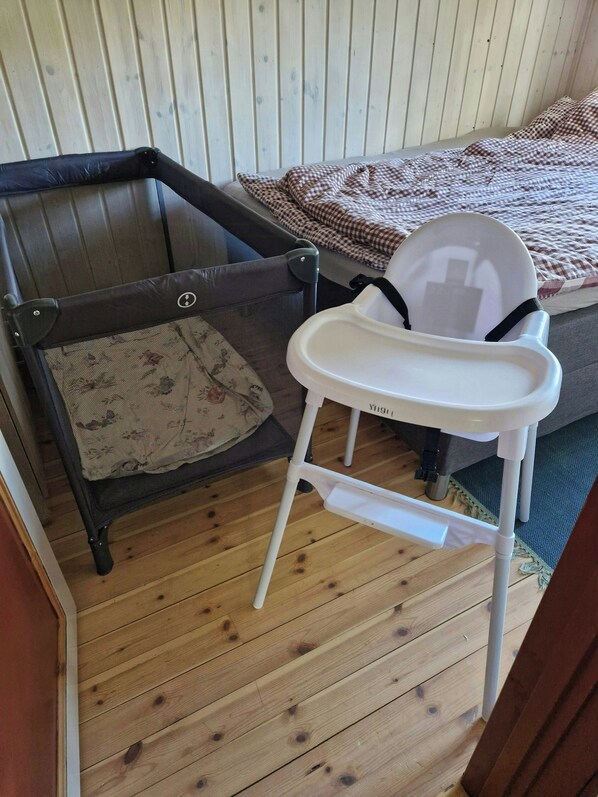A room with a wooden floor, a bed with a checkered blanket, a white baby high chair, and a dark brown sofa with a floral cushion.