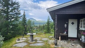 Ein rustikales Blockhaus mit einem Kiesweg, der zu einer Holztür führt, umgeben von viel Grün und Bergen im Hintergrund.